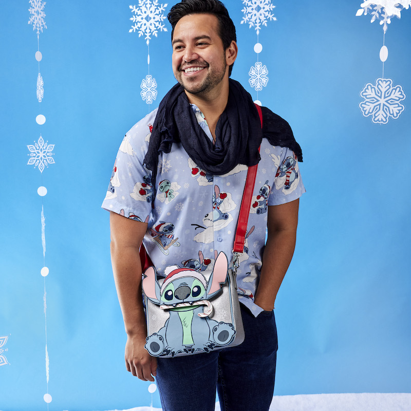 Image of man wearing our Stitch Holiday Unisex Camp Shirt with a black scarf and a holiday Stitch crossbody bag against a blue background with snowflakes.